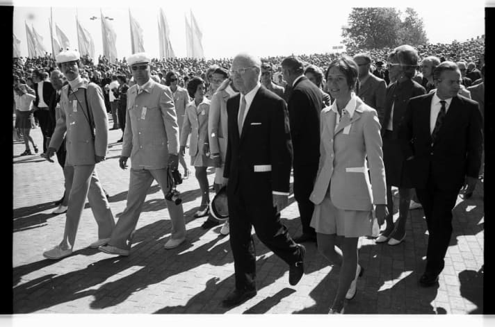   Des visiteurs de marque à la cérémonie d'ouverture de la régate olympique de 1972 : Avery Brundage, président du Comité international olympique (CIO, devant), Willy Daume, président du Comité national olympique (CNO, à droite) ainsi que le président du comité de voile du CNO, Berthold Beitz (derrière Brundage, à l'opposé) flânent parmi la foule sur le parvis du port.