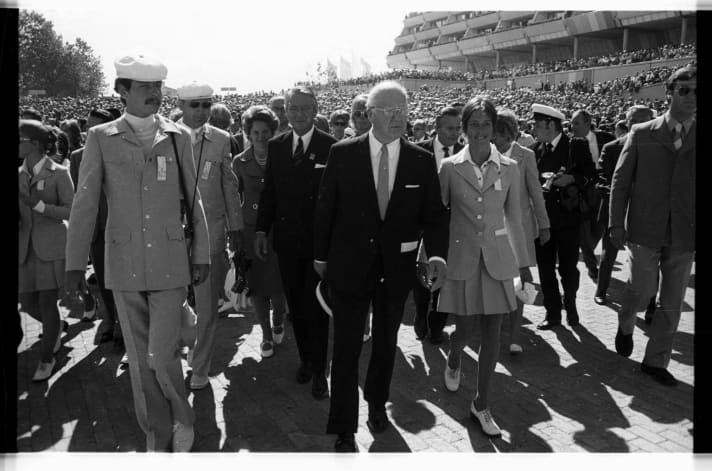   Eröffnungsfeier der Segelolympiade 1972 in Schilksee: Avery Brundage, Präsident des Internationalen Olympischen Komitees (IOC, vorn), Willi Daume, Präsident des Nationalen Olympischen Komitees (NOK, rechts dahinter), sowie der Vorsitzende des Segelausschusses des NOK, Berthold Beitz (hinter Brundage, links), flanieren in Begleitung weiblicher und männlicher Hostessen durch die Menge auf dem Hafenvorfeld. Foto: Friedrich Magnussen (1914-1987), Gesellschaft für Kieler Stadtgeschichte, aus dem Stadtarchiv Kiel. 