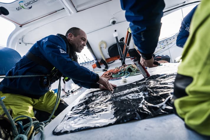 Kevin Escoffier avec une spatule et tout son corps en train de préparer une réparation sur le foc de travail : du sika jusqu'au bord, en dessous un morceau de membrane. Si la colle peut bien prendre et que le support est propre, cela tiendra jusqu'à l'arrivée.