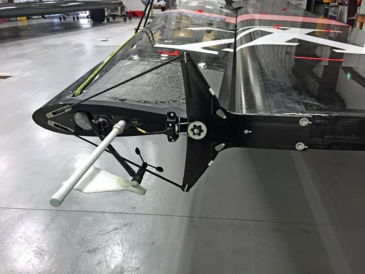   Das Foto zeigt das Bedienelement der Klappe im Topp-Bereich an einem Prototypenflügel von Oracle Team USA. An den Auslegerarmen sind jeweils Trimmleinen befestigt, die durch den Mast nach unten geführt wurden und dort an Hydraulikzylindern enden. Angenommen, mittels des Hydraulikzylinders würde die Leine am oberen Auslegerarm gespannt, so würde sich das Klappenelement nach oben bewegen. Wenn alle Bedienarme um denselben Winkel verstellt werden, entsteht nur ein Profil, kein Twist. Wenn von unten nach oben verschiedene Winkel vorgegeben werden, twistet die Achterkante. Die Bedienelemente wurden zuletzt stark miniaturisiert und verschwinden jetzt im Flügel