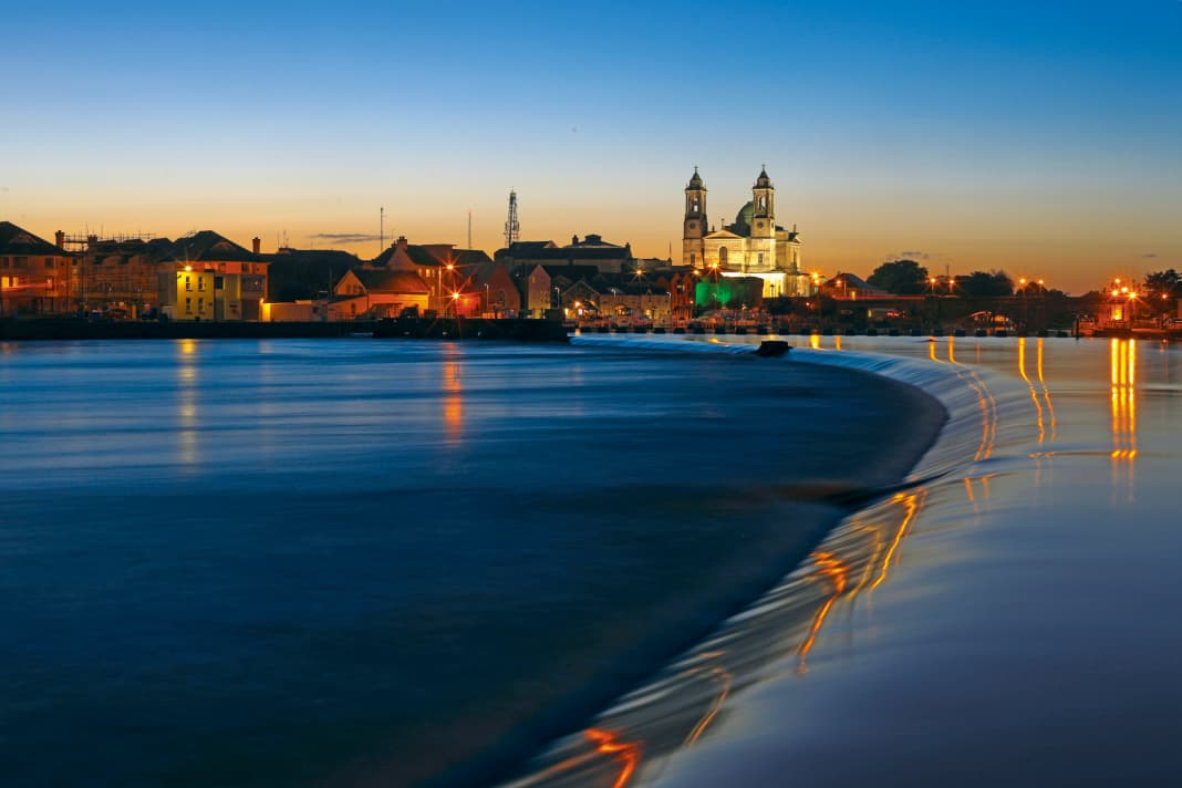 Blick entlang Wehrstufe von Athlone auf Sportbootanleger und  die hell erleuchtete Kathedrale dahinter. Die Langzeitbelichtung täuscht: Der Fluss ist an dieser Stelle alles andere als friedlich