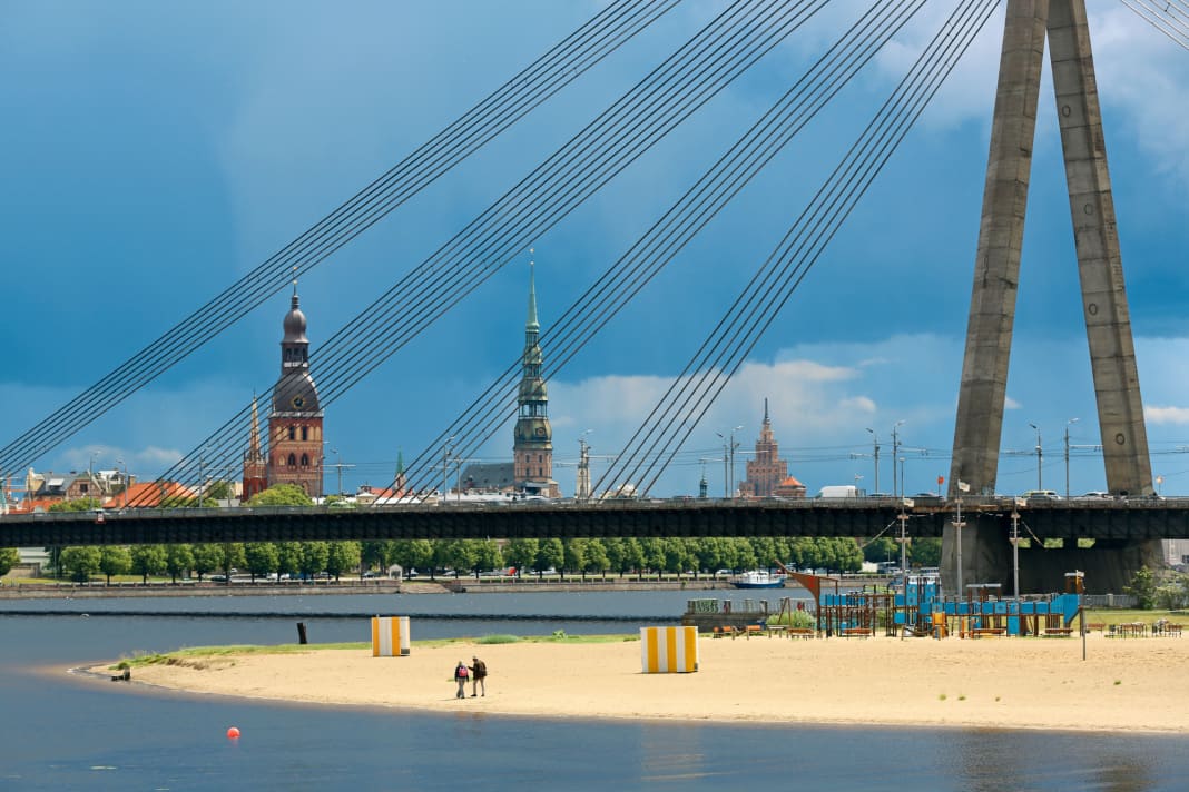 Blick über die Düna auf das Zentrum Rigas, gesehen von der Marina. Im Vordergrund der Kipsala-Strand, dahinter die Vanšu-Brücke. Im Hintergrund von links die Türme des Doms, der Petrikirche und der Akademie der Wissenschaften, eines Baus aus Sowjetzeiten