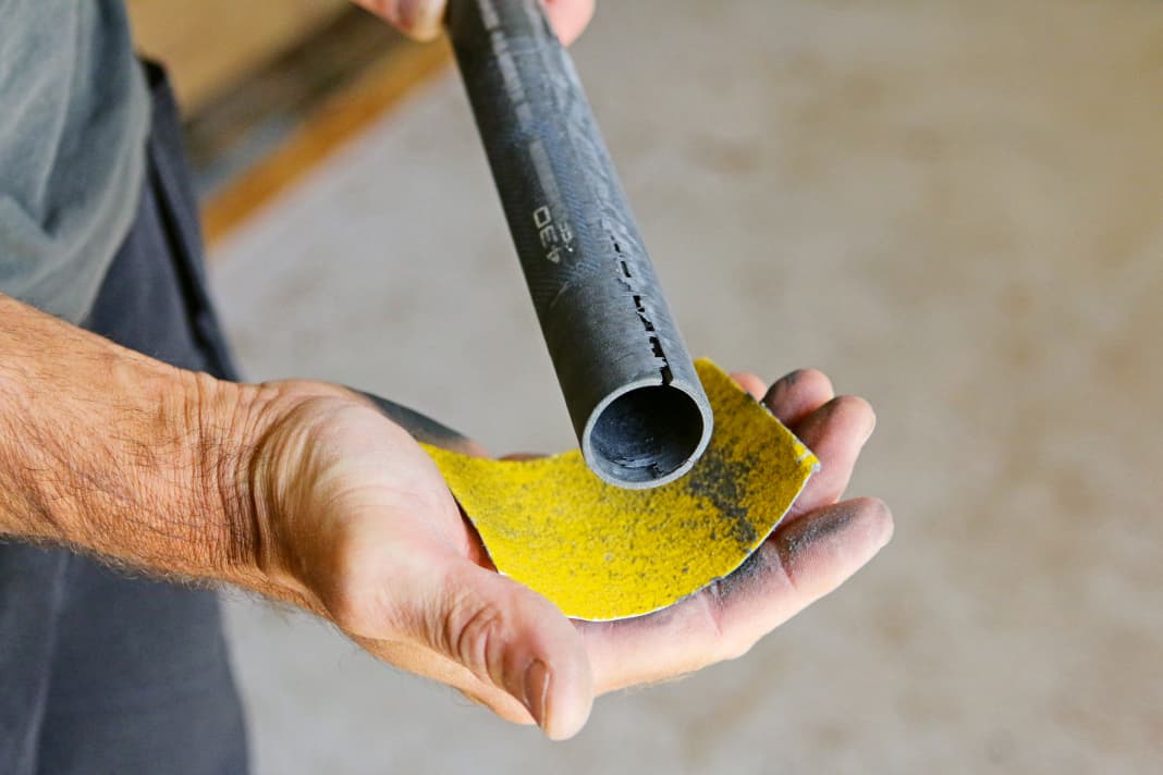 1. Anschleifen: Das Mastoberteil im Bild ist gebrochen, weil der Mast nicht richtig zusammengesteckt war – der Klassiker! Zum Absägen ist der Riss mit 15 Zentimetern zu lang. Auch hier gilt es zunächst, den Reparaturbereich zu überblicken. Stehen auch innen Carbonsplitter ab, sollte man auch eine Kombination der beiden Reparaturen in Betracht ziehen – den schlimmsten Schaden absägen, den Rest, wie im Folgenden erklärt, reparieren. Stickerreste und Carbonsplitter schleift Oli vorsichtig mit Schleifpapier runter. Dazu am besten das Schleifpapier flächig um den Mast legen und diesen in der Hand drehen. Im Zweifel Arbeitshandschuhe tragen, Carbonsplitter unter der Haut sind kein Spaß!