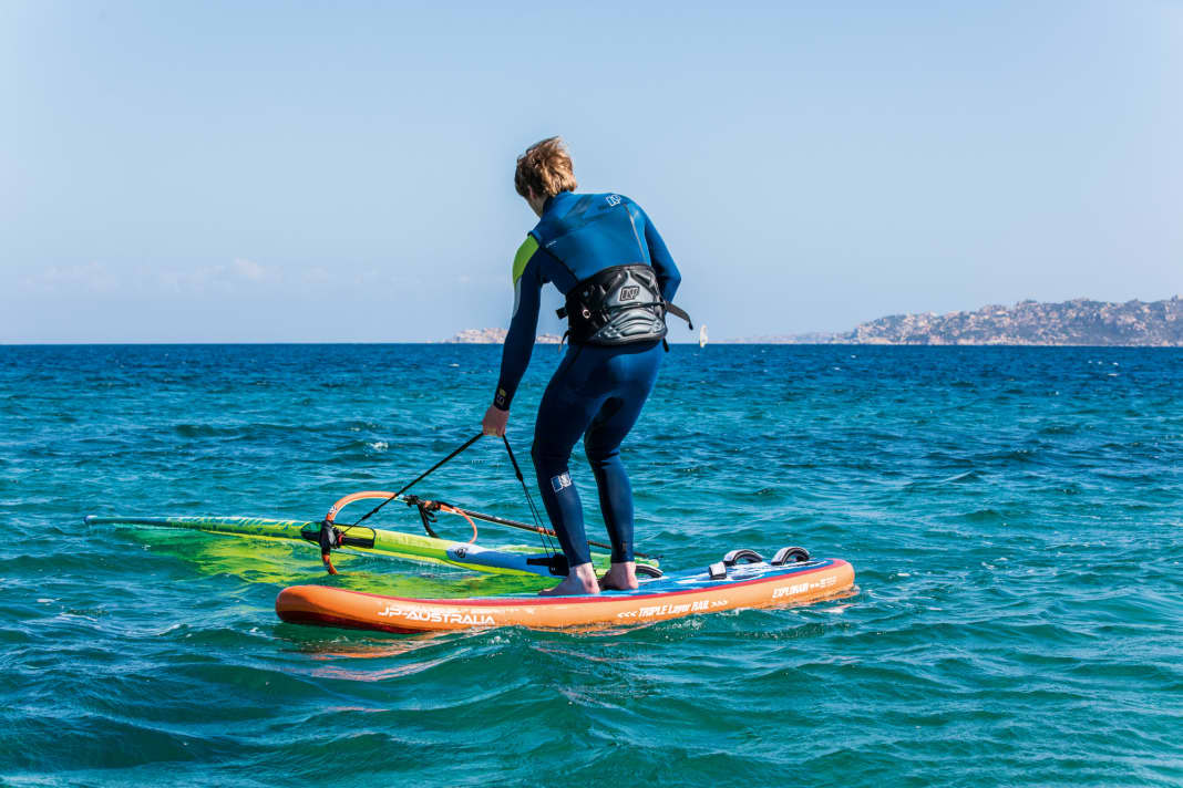 To get started, the board must first be turned into the correct position. To do this, stand on the board with your feet immediately to the left and right of the foot of the mast and grab the furling line far down.