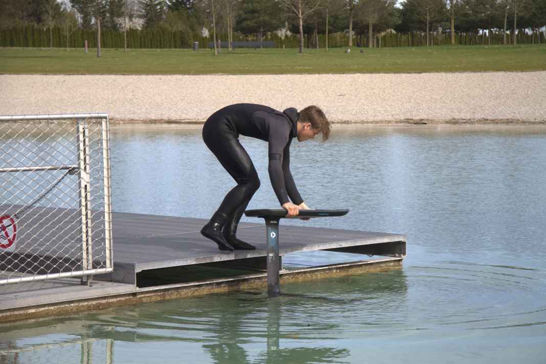Man startet vom Steg aus gebückter Position. Brett und Foil befinden sich in Fahrtposition neben dem Steg. Die Hände halten das Board an den Rails fest. Die eine Hand stabilisiert das Board weiter vorne und die andere näher am Heck. So bleibt das Brett austariert, um es beim Anlaufen sauber neben dem Steg herführen zu können. Der Frontflügel sollte sich dabei nur wenige Zentimeter unter der Wasseroberfläche befinden.