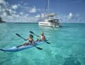 Un couple explore les eaux cristallines en kayak avec pour toile de fond un catamaran à voile en Polynésie française.