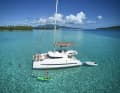 Un catamaran jette l'ancre dans un atoll turquoise sous un ciel d'un bleu éclatant, tandis que les hôtes profitent de l'environnement en faisant du stand-up paddle et en nageant.