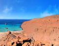 La photo panoramique montre une baie d'un bleu profond, entourée de falaises rocheuses rougeâtres, avec des bateaux isolés sous un vaste horizon.