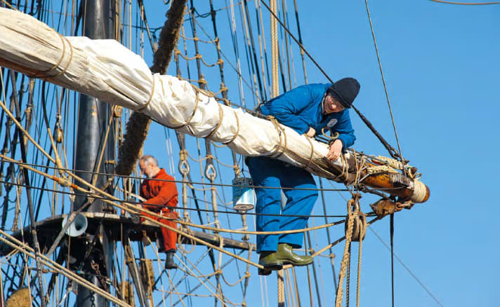 Zwei Crewmitglieder arbeiten hoch oben in den Rahen eines Traditionsschiffes an den Segeln
