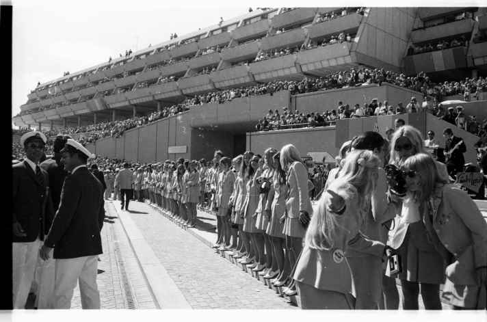   Die Hostessen säumen den Weg für die Ehrengäste. Dahinter das voll besetzte Olympiazentrum Schilksee. Foto: Friedrich Magnussen (1914-1987), Gesellschaft für Kieler Stadtgeschichte, aus dem Stadtarchiv Kiel. 