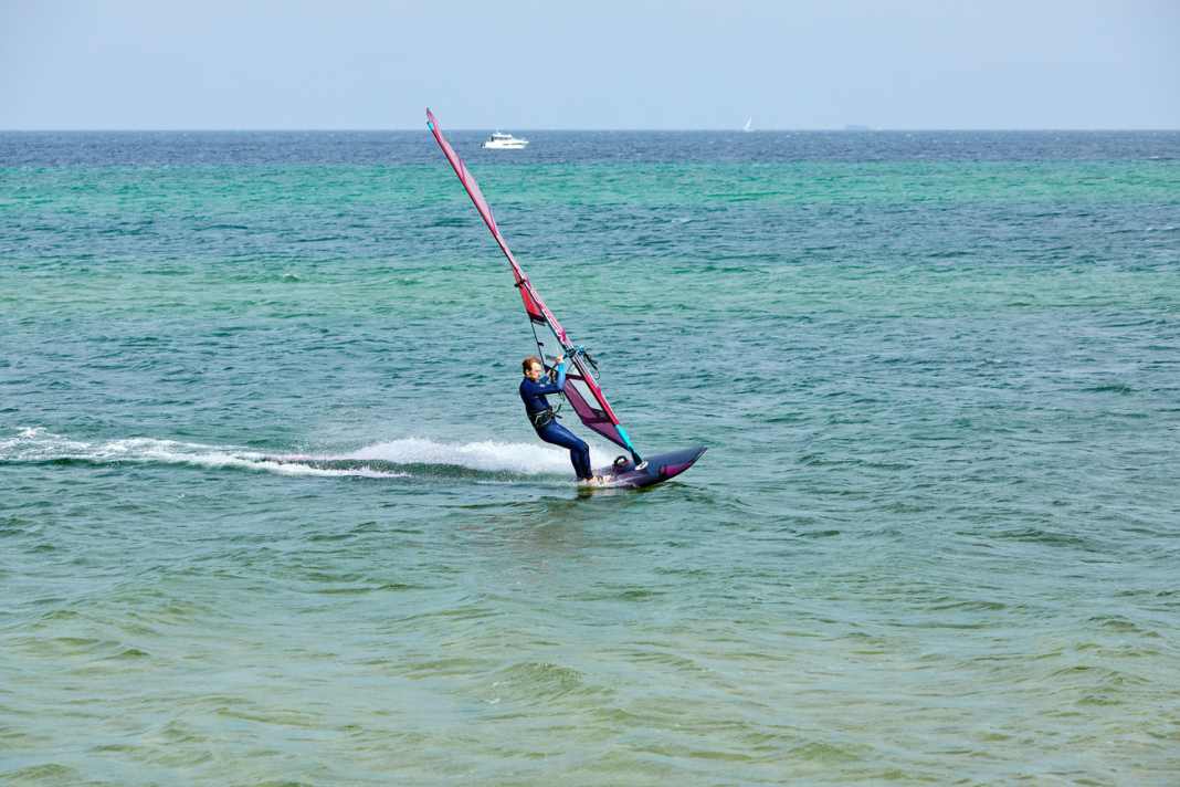 Unhook yourself from the harness in full glide and place your back foot out of the loop onto the windward edge of the board. Place your front foot out of the loop directly next to the foot of the mast.