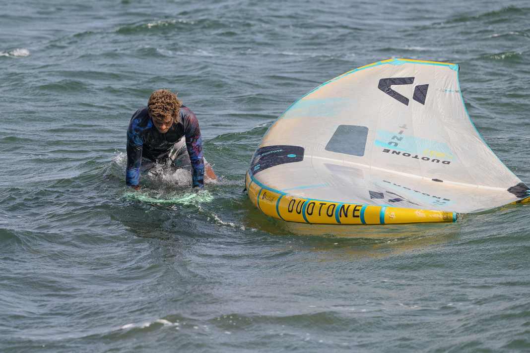 In preparation, Wesley first grabs the wing's handleash and pulls it very close to the board. He then grips the board by the edges and presses it under his body. As always, the wing lies on the downwind side of the board (leeward)