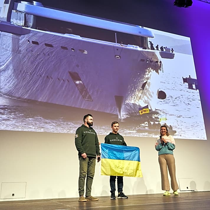 Die Seamaster-Ehrung mit Laudatorin Anastasiya Winkel aus dem Olympiakader des German Sailing Team, Opti-Steuermann Sviatoslav Madonich und Pavlo Dontsov. Im Hintergrund zeigt ein Ausschnitt aus dem bewegenden Film, den Nils Günter für die Seamaster-Ehrung produziert hat, die Protestaktion der Opti-Gruppe gegen die Abramovich-Megayacht “Solaris”