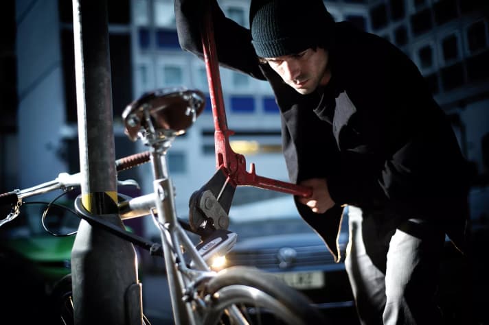 Un homme applique un coupe-boulons sur un antivol à étrier qui sert à sécuriser un vélo sur un poteau métallique.