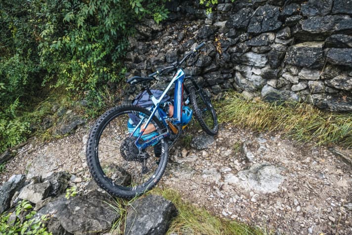   Drehen Sie jetzt die Ihnen zugewandte Kurbel so, dass das Pedal unten sitzt. Machen Sie sich jetzt so klein wie möglich und packen Sie das Bike an Gabel und Kurbel. Strecken Sie Ihren Kopf so weit wie möglich unter das Unterrohr. Jetzt braucht man nur das Bike etwas zu lupfen und gleichzeitig aufzustehen (2).