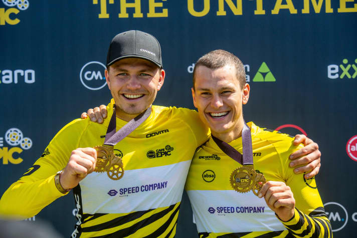 A historic moment for the German mountain bike scene: Georg Egger and Lukas Baum pose as overall winners of the Cape Epic 2022 with a medal in the yellow jersey.