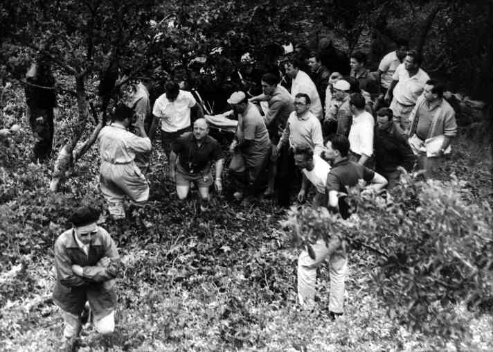 Coaches, helpers, reporters and photographers surround the fallen racer, who is rescued from the ravine on a stretcher