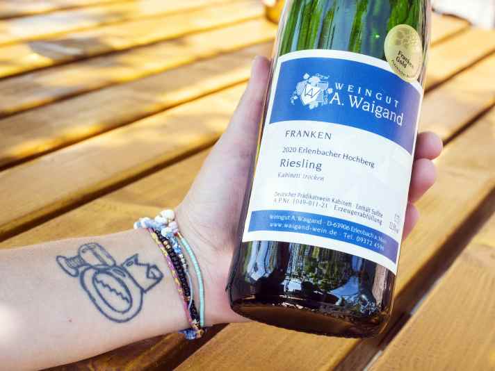 Der Beruf geht unter die Haut: Winzerin Verena Waigand präsentiert stolz ihren prämierten Wein und ihr Tattoo mit traditionellem Bocksbeutel und Römer Weinglas.