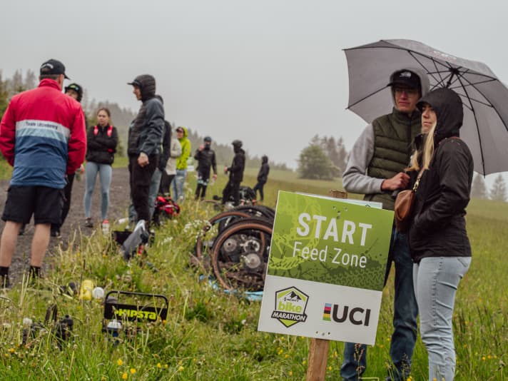   Bei acht Grad und Nieselregen mussten sich die Marathon-Teilnehmer heute durch die Hügellandschaft rund um Willingen kämpfen. Doch wenn die Muskeln so richtig am Feuern sind, bleibt wenigsten der Körper auf Betriebstemperatur. Die Helfer an der Feed Zone am Anstieg zur dritten Runde, mussten sich mit Daunen-Jacke und Regenschirm gegen das raue Wetter hier im Upland schützen.