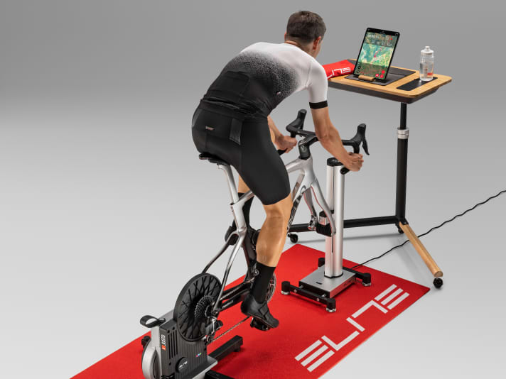 So kann ein Indoor-Cycling-Setup aussehen: Elite Justo und Rizer auf Matte mit Tisch und Tablet für virtuelle Fahrten.