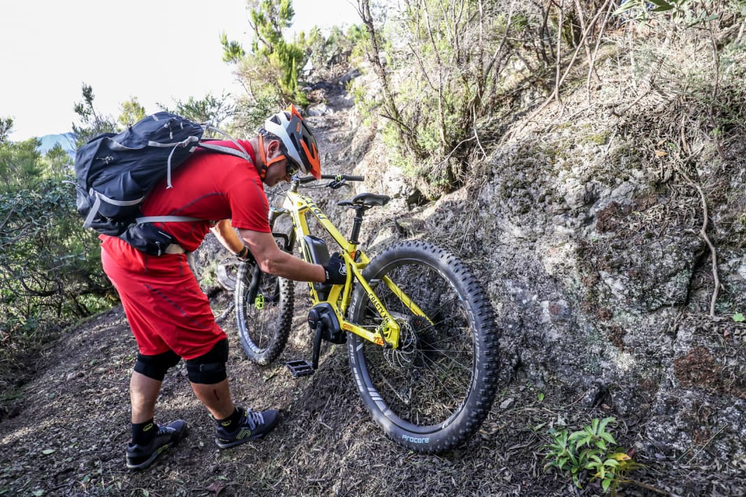 Damit das E-MTB bequemer auf dem Rücken sitzt, stellen Sie die Gurte des Rucksacks so eng ein, dass dieser möglichst nah an den Schultern sitzt. Die Kurbel mit Pedal zeigt nach unten. Greifen Sie jetzt das Bike am Gabelholm und irgendwo im Bereich der Mitte des Sitzrohrs (Bild 1).