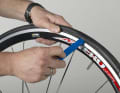 2. slide a tyre lever next to the valve under the tyre foot. Work on the opposite side of the sprocket on the rear wheel to avoid injury if you slip off. The blunt side of the lever points towards the inner tube. Lever the tyre over the side of the rim before placing the second lever about ten centimetres away. Here too, lever the side of the tyre over the braking surface.