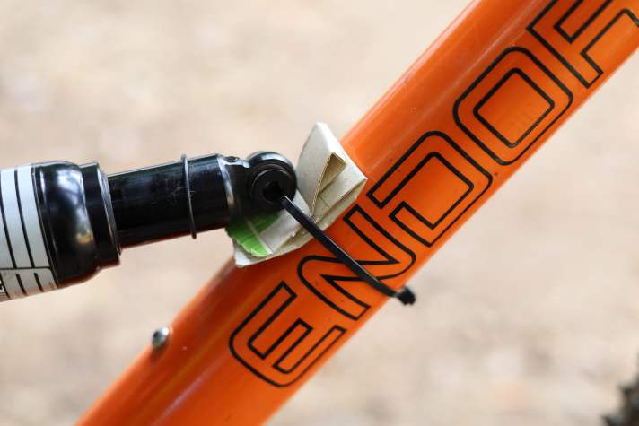 Da sich der Dämpfer nicht mehr ausbauen lässt, muss er anderweitig fixiert werden, ohne die maximale Kompression des Hinterbaus zu blockieren. Ein Kabelbinder und ein Stück Pappe halten ihn an Ort und Stelle, ohne dass der Rahmen weiter beschädigt wird.