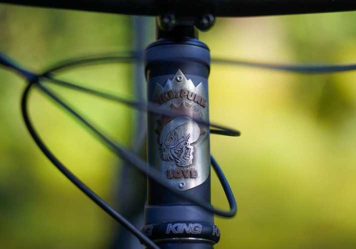   Obwohl die originale Steuerrohr-Plakette mit dem Schriftzug "Patria – Seit 1898" lässig aussieht, sollte ein Kunstwerk von Jen Green ans Bike. Nach einer handgemalten Vorlage von Henri fertigte die amerikanische Headbadge-Designerin diese Plakette. Das Motiv symbolisiert die drei Säulen bzw. Konstanten in Henri's Leben: Bike, Punk, Liebe. Kosten: 160 Euro. 