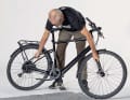So drehen Sie ein schweres E-Bike rückenschonend um: Beugen Sie sich über den Rahmen. Greifen Sie außen auf etwa halber Länge an Sitzstrebe und Gabelholm.