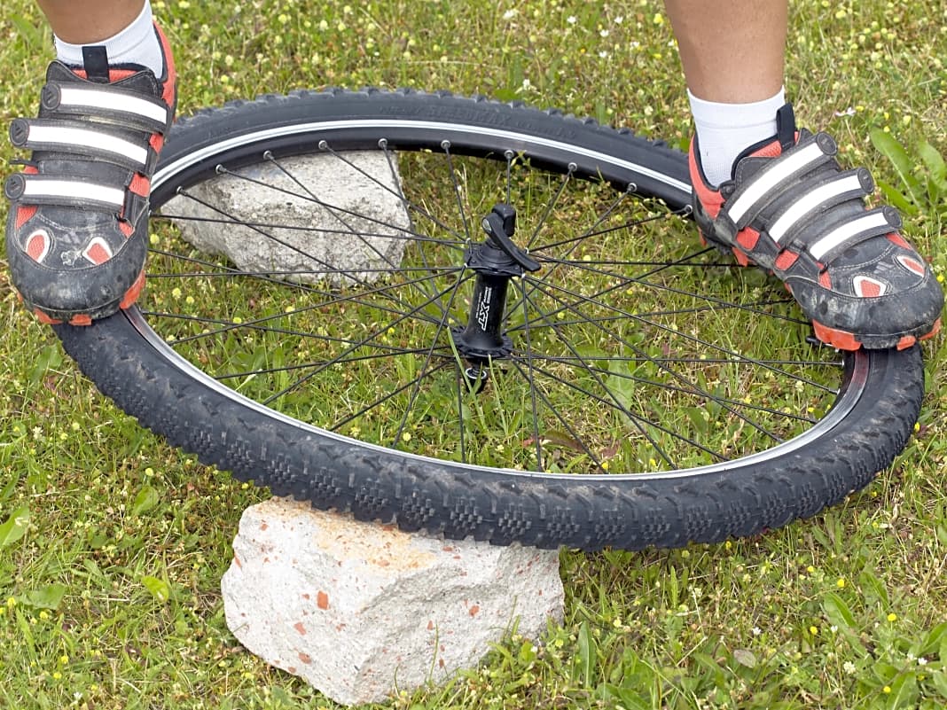 KAPITALER ACHTER

Das Laufrad verzieht sich nicht nur durch gebrochene Speichen. Durch extreme seitliche Belastungen oder einen Einschlag mit hoher Geschwindigkeit kann das Laufrad “chippen”. Um die Tour trotzdem fortsetzen zu können, sollten Sie den “Achter” aus dem Laufrad drücken. Legen Sie das Laufrad dazu auf etwas Festes, zum Beispiel Steine wie im Bild. Die Stellen, an denen die Felge knickt, sollten aufliegen. Stellen Sie sich mit einem Fuß auf die gerade Seite der Felge. Mit dem anderen Fuß belasten Sie die verbogene Seite. Kontrollieren Sie den Erfolg und weiderholen Sie den Vorgang falls nötig. Tipp: Geht am besten zu zweit. Eventuell müssen Sie für die Weiterfahrt die Felgenbremse aushängen, aber zumindest passt das Laufrad durch die Gabel. Zuhause von einem Fachmann kontrollieren lassen.