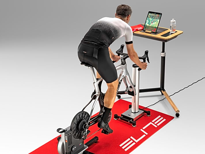 So kann ein Indoor-Cycling-Setup aussehen: Elite Justo und Rizer auf Matte mit Tisch und Tablet für virtuelle Fahrten.